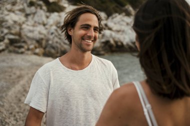 Beyaz erkek, kumsalda yürürken kız arkadaşına gülümsüyor.