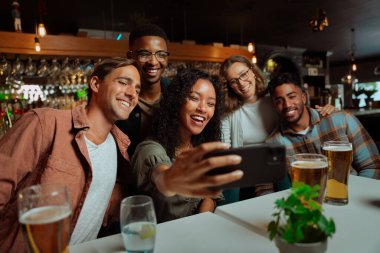 Bir grup yetişkin arkadaş restoranda yemek yiyor ve selfie çekiyorlar.