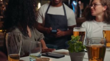 african american waiter holding card machine while friends pay for drinks with cellphone