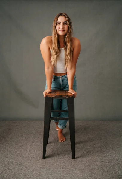 Young female leaning on a high stool with her hands on the stool in her jeans and t shirt bare foot High quality photo