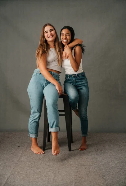 Duas Amigas Sorrindo Vestindo Jeans Tops Brancos Compartilhando Banquinho Estúdio — Fotografia de Stock