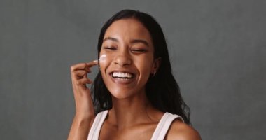 Genç, melez bir kadın yüzüne krem sürüyor ve yüz kremini nazikçe yüzüne sürüyor.