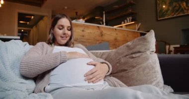 Heavily pregnant woman sitting on her sofa gently rubbing her tummy and talking to the unborn baby. High quality 4k footage
