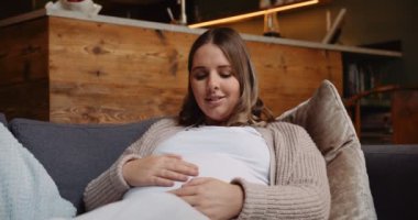 Pregnant female lying on the sofa and gently rubbing her tummy in her apartment. High quality 4k footage