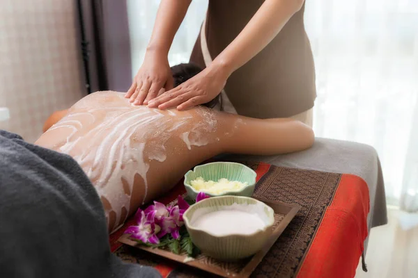 Spa therapist applying scrub salt and cream on young woman back at salon spa. Hands massaging female back with scrub.