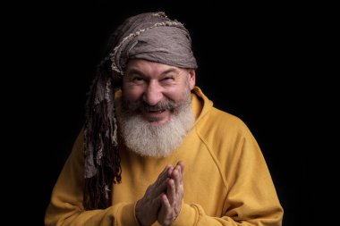 Olgun sakallı beyaz adam kameraya sinsi sinsi bakar ve gülümser, gri eşarp türbanı, siyah arka plan