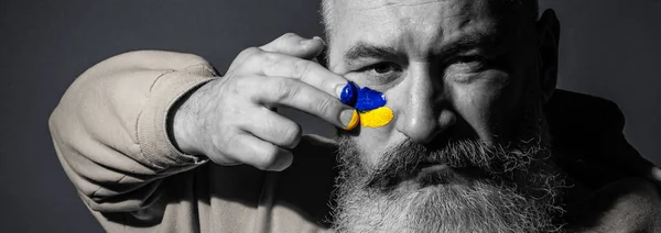 Mature bearded man dressed yellow hoodie draws Ukrainian flag on his face, protest against war, black and white portrait