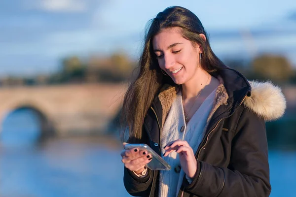 Latynoska młoda kobieta używa smartfona do portali społecznościowych w pobliżu rzeki Tajo, Toledo. Zimowy styl zimowy — Zdjęcie stockowe