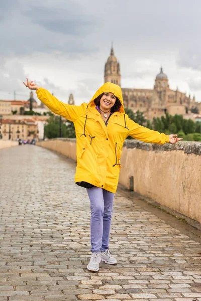 famous place cathedral monument. happy young hispanic woman wearing a yellow waterproof coat. positive person in front of historical architecture of camino de santiago