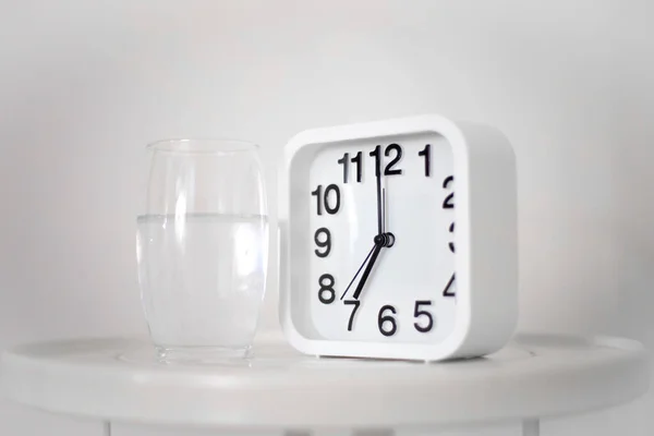White alarm clock and glass of water on table in bedroom. Wake up early in the morning by the alarm bell and drink water.