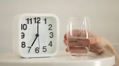Yatak odasındaki masada beyaz çalar saat ve bir bardak su. El ele tutuşan cam, yakın görüş. Sabah erken kalk ve su iç. Sağlıklı yaşam için iyi alışkanlıklar, susuzluk kavramı