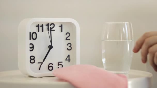 Λευκό ξυπνητήρι και ένα ποτήρι νερό στο τραπέζι στην κρεβατοκάμαρα. Γυαλί χειρός, κοντινή θέα. Ξυπνήστε νωρίς το πρωί και πιείτε νερό. Καλές συνήθειες για υγιή τρόπο ζωής, έννοια αφυδάτωσης — Αρχείο Βίντεο