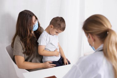 Küçük oğluyla birlikte genç esmer kadın çocuk doktorunda danışmanlık yapıyor. Çocuğun karın ağrısı var. Doktor, çocuk ve anne koronavirüs ve grip salgını sırasında maske takıyor. Covid 19.