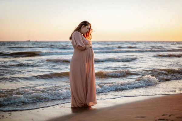 Молодая беременная женщина с прекрасным видом на море на заднем плане. Счастливая и спокойная беременная женщина с длинными волосами и розовым платьем стоит на пляже. Романтический вид, океан, закат, материнство. — стоковое фото