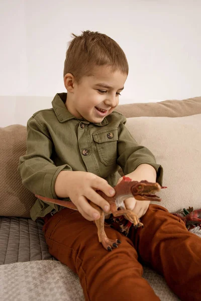 Pequeño y lindo chico caucásico jugando con dinosaurios en casa. Interior y ropa en colores tierra natural. Ambiente acogedor. Niño divirtiéndose con juguetes. — Foto de Stock