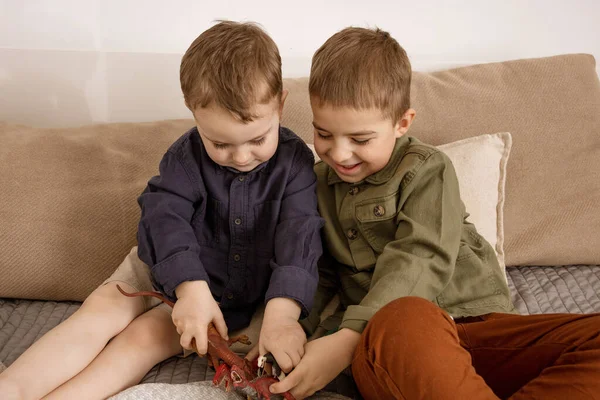 Dos pequeños y lindos chicos caucásicos jugando con dinosaurios en casa. Interior y ropa en colores tierra natural. Ambiente acogedor. Niños divirtiéndose con juguetes, dos amigos. — Foto de Stock