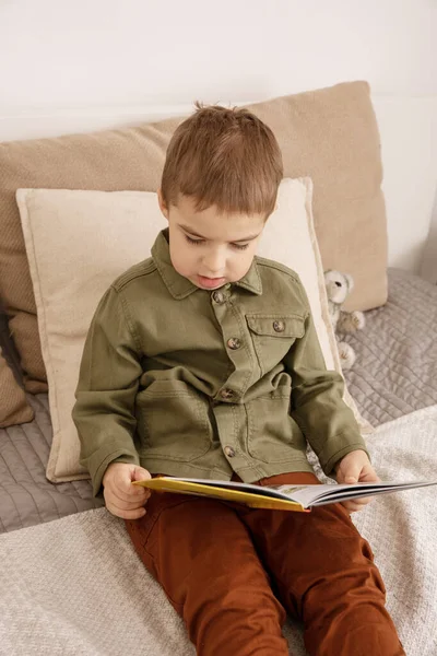 Pequeño y lindo chico caucásico leyendo un libro en la cama en casa. Interior y ropa en colores tierra natural. Ambiente acogedor. Niño lee un cuento de hadas. — Foto de Stock