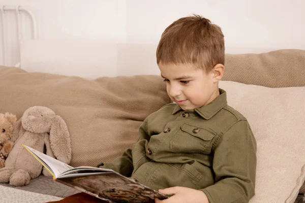 Pequeño y lindo chico caucásico leyendo un libro en la cama en casa. Interior y ropa en colores tierra natural. Ambiente acogedor. Niño lee un cuento de hadas. — Foto de Stock