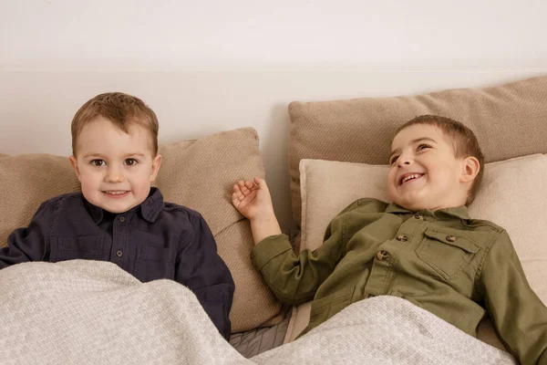 Two little and cute caucasian boys playing together on the bed at home. Interior and clothes in natural earth colors. Cozy environment. Children having fun, two brothers. — Foto de Stock