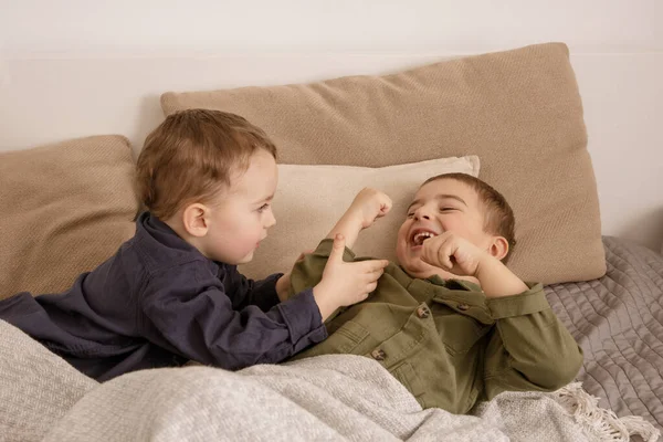 Two little and cute caucasian boys playing together on the bed at home. Interior and clothes in natural earth colors. Cozy environment. Children having fun, two brothers kittle each other. — Foto de Stock