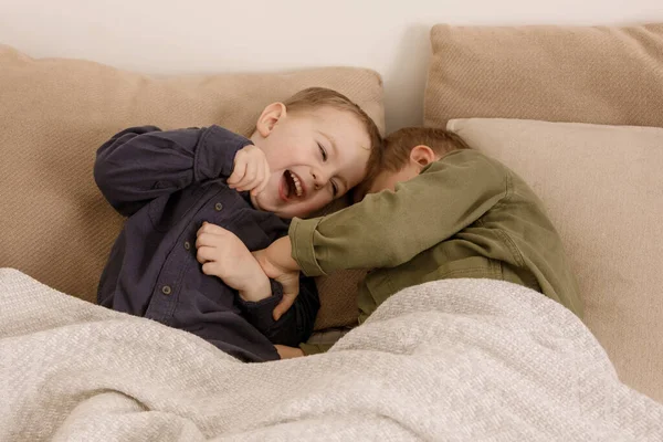 Two little and cute caucasian boys playing together on the bed at home. Interior and clothes in natural earth colors. Cozy environment. Children having fun, two brothers kittle each other. — Foto de Stock