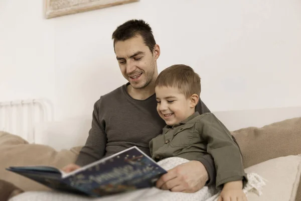 Father and son reading a book on the bed at home. Young attractive man and little boy resting in bedroom. Natural earth colors. Cozy environment. Father reads a fairy tale for his child. — Stock Photo, Image