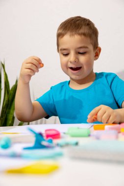 Mavi gömlekli küçük çocuk masasında el işi yapıyor. Çocuk renkli kağıda resim çizer, evde oynar.