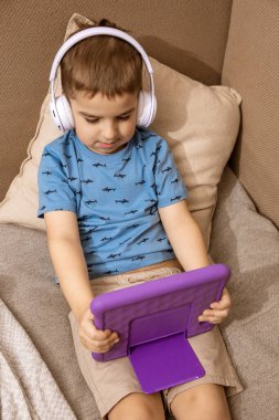 Beyaz kulaklıklı, mavi gömlekli beyaz çocuk evde mor dijital tabletle çizgi film izliyor. Modern çocuk ve eğitim teknolojisi.