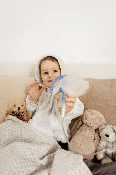 Sick little boy with inhaler for cough treatment. Unwell kid doing inhalation on his bed. Flu season. Medical procedure at home. Interior and clothes in natural earth colors. Cozy environment. — Foto de Stock