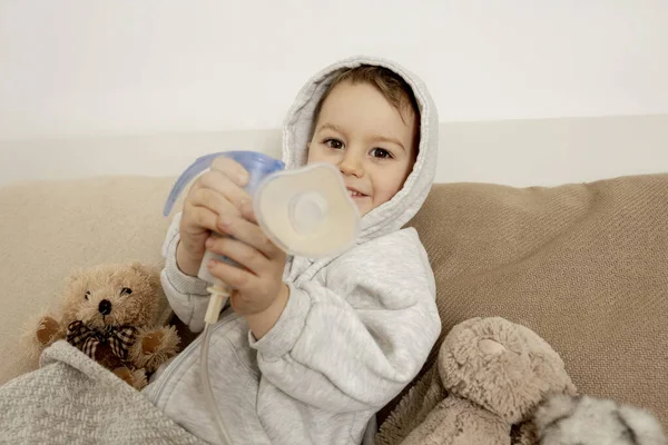 Sick little boy with inhaler for cough treatment. Unwell kid doing inhalation on his bed. Flu season. Medical procedure at home. Interior and clothes in natural earth colors. Cozy environment. — Foto de Stock