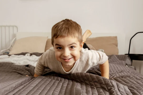 Portrait of little, smiling caucasian boy on the bed at home. Cute child relaxing, resting in bedroom. Positive emotions. Cozy and modern interior. Natural, earth colors. Casual clothes. — Foto de Stock