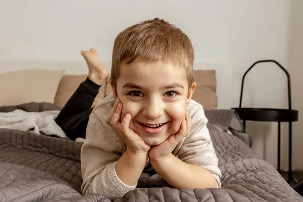 Portrait of little, smiling caucasian boy on the bed at home. Cute child relaxing, resting in bedroom. Positive emotions. Cozy and modern interior. Natural, earth colors. Casual clothes. — Foto de Stock