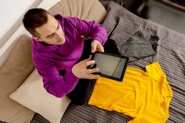 Man with violet hoodie using digital tablet, takes a photo of his old clothes to sell them online. Selling on website, e-commerce. Reuse, second-hand concept. Conscious consumer, sustainable lifestyle — Stock fotografie