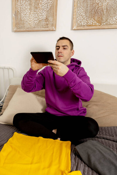 Man with violet hoodie using digital tablet, takes a photo of his old clothes to sell them online. Selling on website, e-commerce. Reuse, second-hand concept. Conscious consumer, sustainable lifestyle