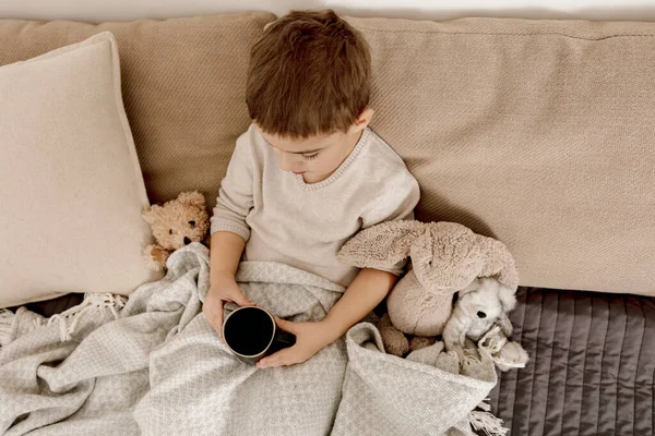 Niño enfermo bebiendo té caliente en la cama en casa. Malestar, niño enfermo envuelto en una manta, con taza en su habitación. Temporada de gripe. Interior y ropa en colores tierra natural. Ambiente acogedor. — Foto de Stock