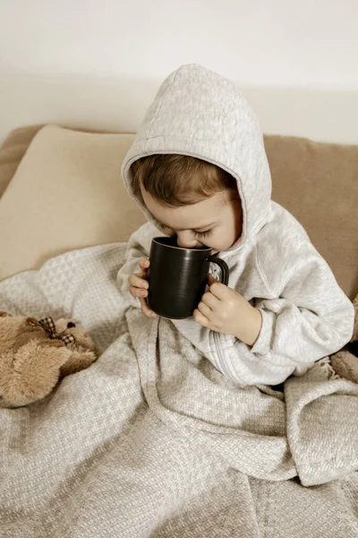 Больной маленький мальчик пьет горячий чай на кровати дома. Больной ребенок, завернутый в одеяло, с кружкой в комнате. Сезон гриппа. Интерьер и одежда в натуральных цветах земли. Уютная обстановка. — стоковое фото