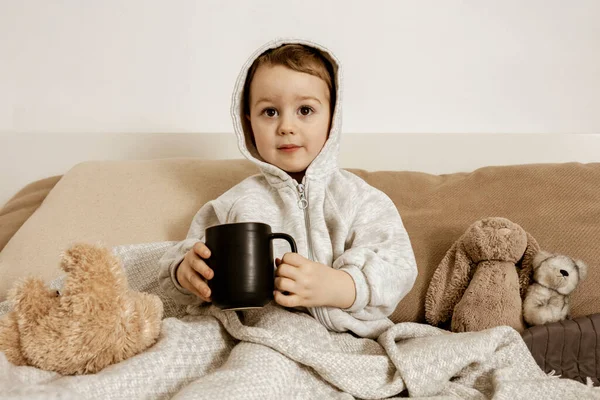 Niño enfermo bebiendo té caliente en la cama en casa. Malestar, niño enfermo envuelto en una manta, con taza en su habitación. Temporada de gripe. Interior y ropa en colores tierra natural. Ambiente acogedor. — Foto de Stock