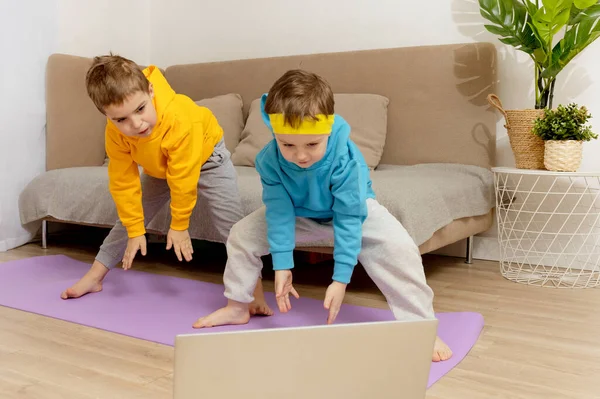Двое белых детишек занимаются дома фитнесом. Дети веселятся во время спорта. Мальчики тренируются, смотрят в компьютер. Спорт, йога онлайн. Тренировка в квартире. Здоровый образ жизни. — стоковое фото