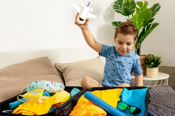 Kleiner kaukasischer Junge mit blauem Hemd bereit für den Urlaub. Glückliches Kind packt Kleidung in einen Reisekoffer. Touristen, Urlaubsfreuden. Kind zu Hause, Vorbereitung auf das Fliegen. Moderner und gemütlicher Innenraum. — Stockfoto