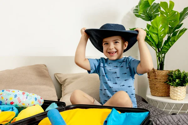 Kleiner kaukasischer Junge mit blauem Hemd bereit für den Urlaub. Glückliches Kind packt Kleidung in einen Reisekoffer. Touristen, Urlaubsfreuden. Kind zu Hause, Vorbereitung auf das Fliegen. Moderner und gemütlicher Innenraum. — Stockfoto