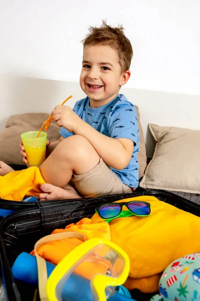 Pequeño chico caucásico con camisa azul listo para las vacaciones. Niño feliz empaca la ropa en una maleta para viajar. Turista, alegría de las vacaciones. Niño en casa, preparándose para volar. Interior moderno y acogedor. — Foto de Stock