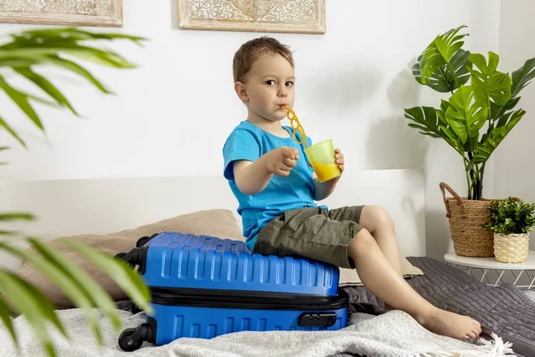 Kleiner kaukasischer Junge mit blauem Hemd bereit für den Urlaub. Glückliches Kind mit blauem Koffer auf Reisen. Touristen, Urlaubsfreuden. Kind zu Hause, Vorbereitung auf das Fliegen. Moderner und gemütlicher Innenraum. — Stockfoto