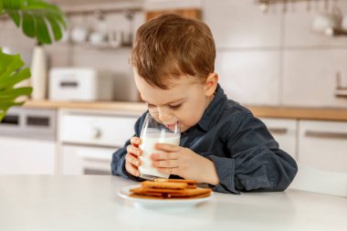 Küçük çocuk mutfakta oturuyor ve süt içiyor. Bardakta taze süt, süt ürünleri, sağlıklı içecek. Sağlık sigortası, kalsiyum kaynağı, laktoz. Konforlu ve modern iç mekan. Günlük kıyafetleriyle anaokulu çocuğu..