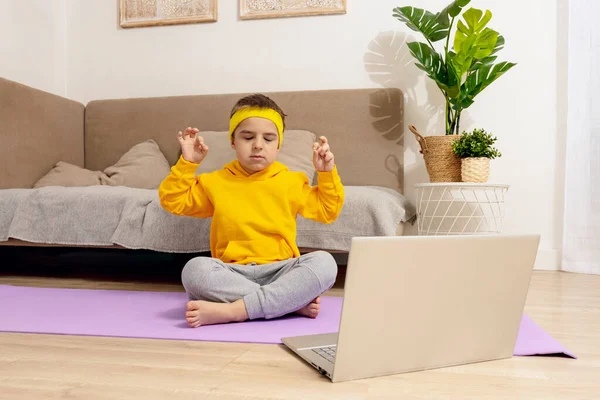 自宅でヨガの練習をしている小さな白人の少年。子供はスポーツ中に楽しんでいます。子供はコンピューターを見て訓練をする。スポーツ、ヨガオンライン。アパートで働いてる。健康的なライフスタイル. — ストック写真