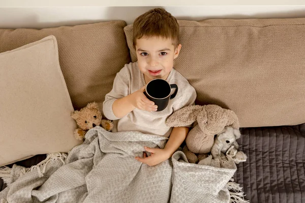 Adorable niño bebiendo té caliente en la cama en casa y relajante. Niño descansando, envuelto en una manta, con taza en su habitación. Interior y ropa en colores tierra natural. Ambiente acogedor. — Foto de Stock