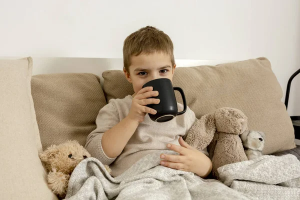 Adorable niño bebiendo té caliente en la cama en casa y relajante. Niño descansando, envuelto en una manta, con taza en su habitación. Interior y ropa en colores tierra natural. Ambiente acogedor. — Foto de Stock