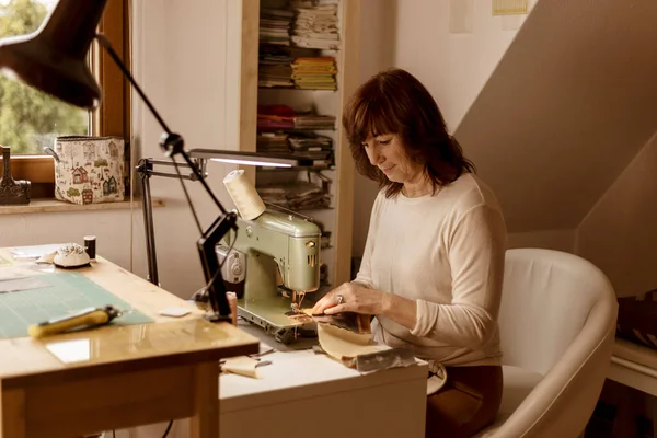 Caucasian brunette woman sewing fabric with a vintage, retro sewing machine. Fashion, creation and tailoring. Process of sewing in atelier or workshop. Special hobby. Cozy interior and warm colors. — Stock Photo, Image