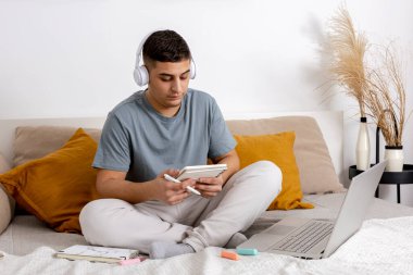 Gündelik giysileri olan genç bir adam evde dizüstü bilgisayarla yatakta oturuyor ve ders çalışıyor. Adam öğretmeniyle video görüşmesi yapmak için e-öğrenme platformunu kullanıyor. Uzaktan eğitim. İçerisi çok rahat..