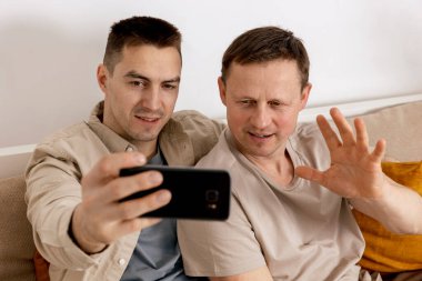 Gündelik kıyafetleri olan mutlu eşcinsel çift ellerinde akıllı telefon tutuyorlar ve arkadaşlarıyla internetten konuşuyorlar. Video görüşmesi yapıyorlar. Homoseksüel ilişkiler ve alternatif aşk.