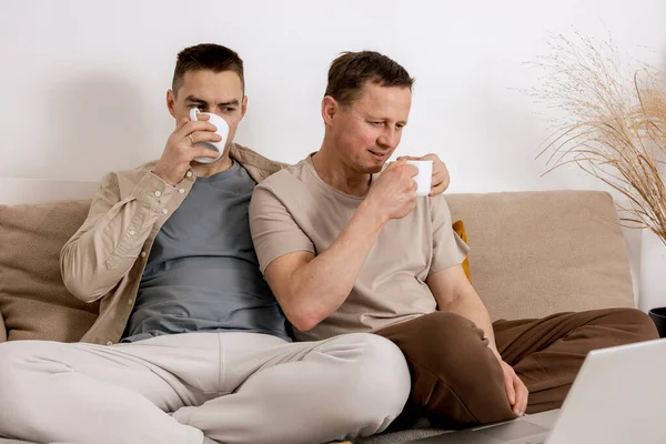 Šťastný gay pár s neformálním oblečením tráví čas doma, dívají se na film na notebooku a pijí čaj. Dva běloši odpočívají. Homosexuální vztahy a alternativní láska. — Stock fotografie